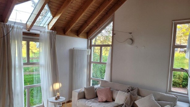 A cozy room with a wooden ceiling, a white sofa with cushions, a round side table, and a large window with curtains. - Holiday Home 'Lichtdurchflutetes Architektenhaus' with Mountain View, Terrace and Wi-Fi (Balingen)