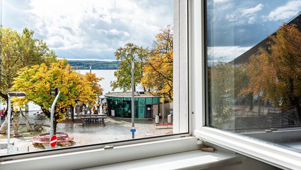 A view through a window of a lakeside area with a cafe, benches, and trees with autumn foliage. - Apartment '2 Mit Seeblick' with Lake View and Wi-Fi (Überlingen)
