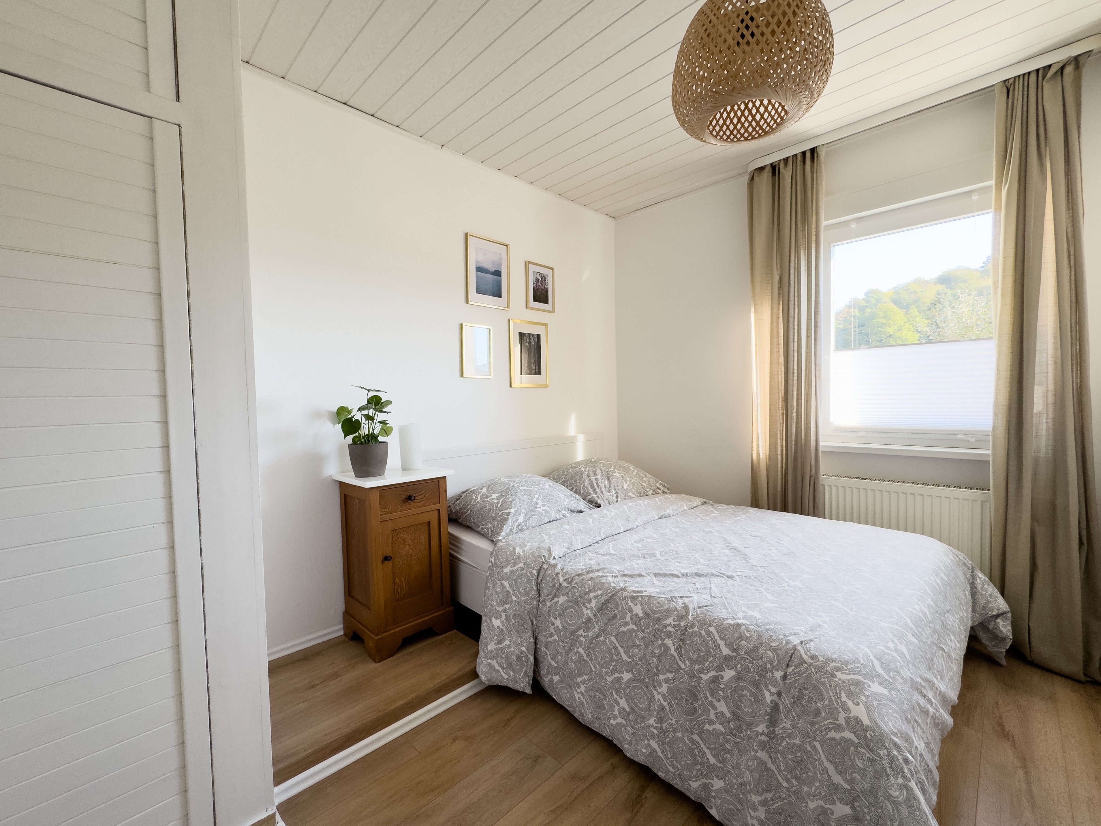 A bedroom with a bed, a nightstand, a potted plant, a window with curtains, and framed pictures on the wall.