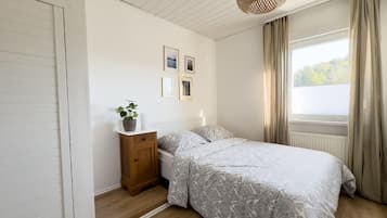 Ein Schlafzimmer mit einem Bett, einem Nachttisch, einer Zimmerpflanze, einem Fenster mit Vorhängen und gerahmten Bildern an der Wand.