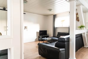 Ein modernes Wohnzimmer mit einem schwarzen Ledersofa, einem kleinen runden Couchtisch, einem Flachbildfernseher und einem Fenster mit Vorhängen.