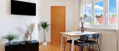 A modern living room with a flat-screen TV, a wooden door, a dining table with chairs, and potted plants.