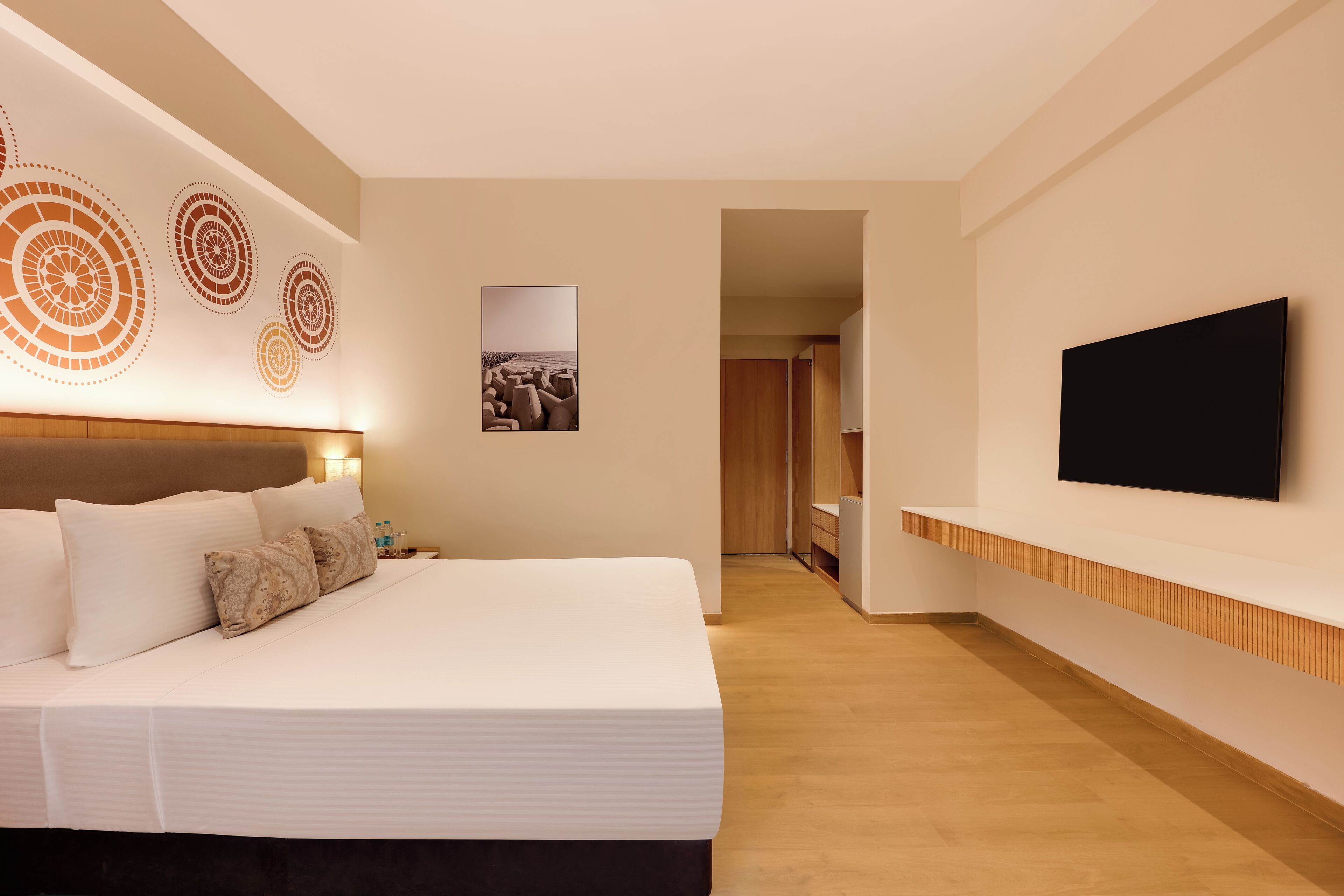 Una habitación de hotel moderna con una cama grande, un televisor de pantalla plana montado en la pared, piso de madera y un cuadro de una playa.