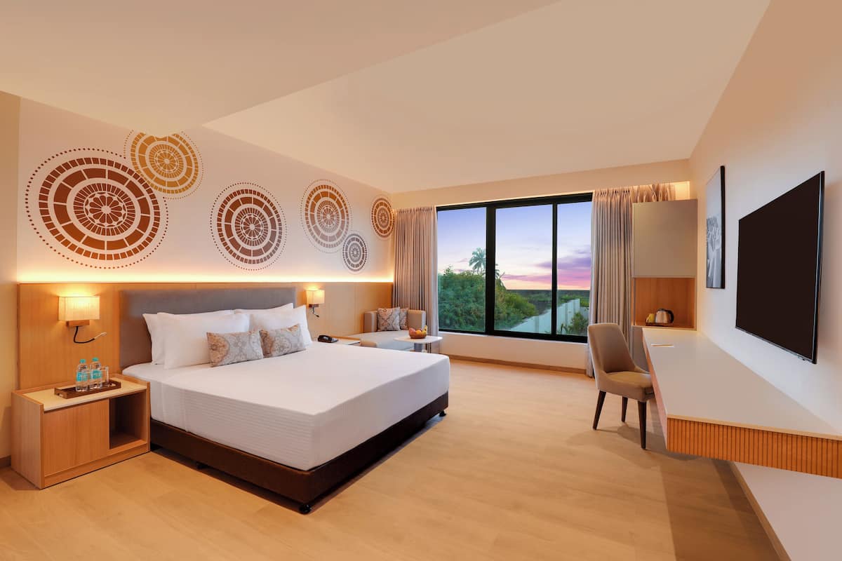 A modern hotel room with a large bed, a desk, a TV, and a large window with a view of the sunset.