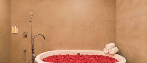A bathroom with a bathtub filled with red bath beads, a showerhead, and a towel rack with towels.