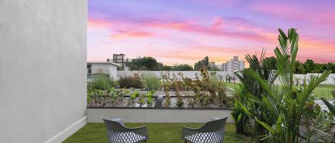 Una terraza en la azotea con dos sillas, un pequeño jardín y vistas a edificios bajo un cielo al atardecer.