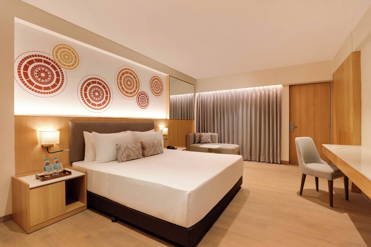 A modern hotel room with a large bed, a desk, a chair, a small table, and a wall-mounted lamp.