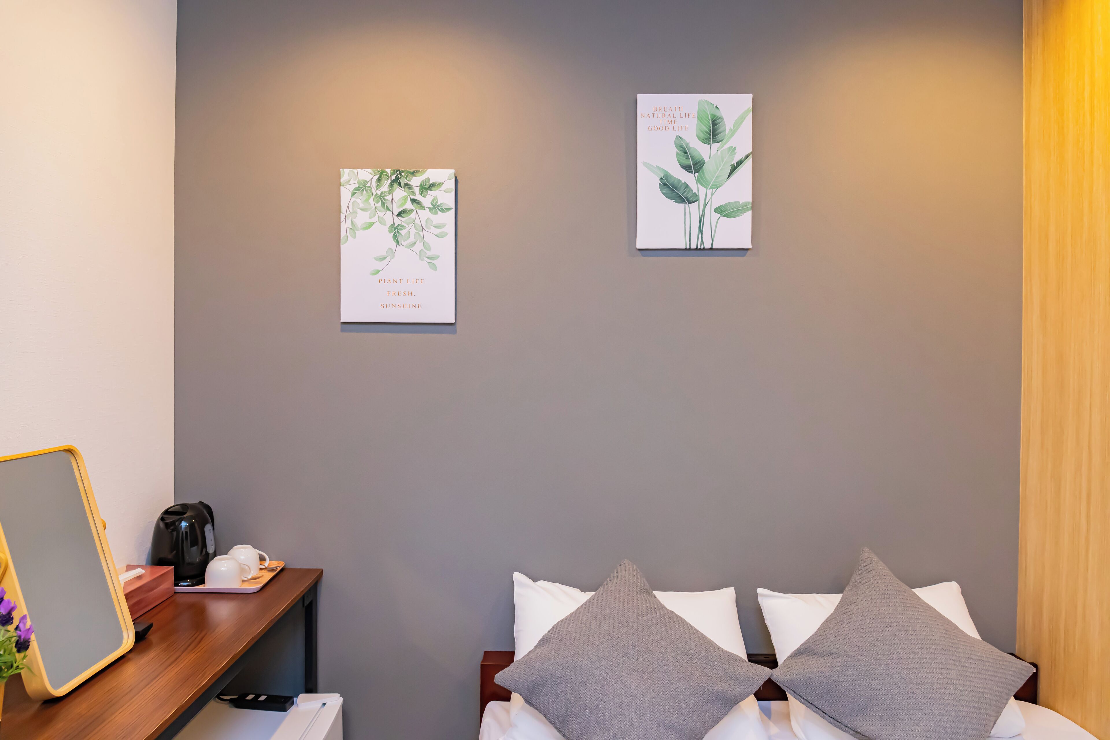 A hotel room with a bed, two pillows, a bedside table with a kettle and cups, and two wall art pieces.
