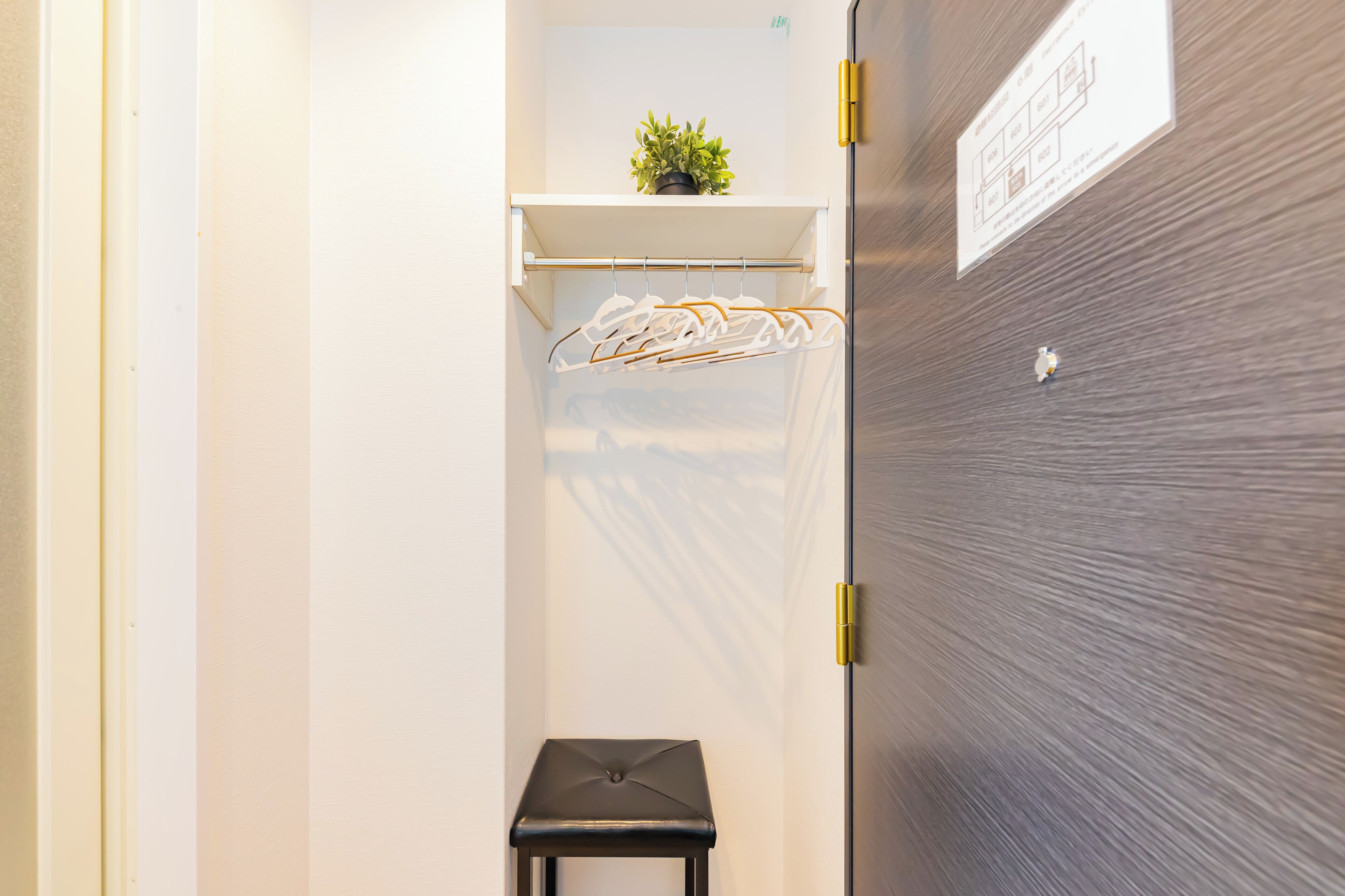A compact room with a small stool, a white shelf with a plant, and a wall-mounted coat rack.