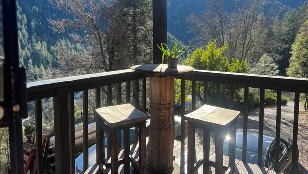 A wooden deck with two square tables, a potted plant, and a view of a mountainous landscape.