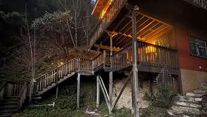 A wooden staircase leading to a balcony with string lights, surrounded by trees and stone steps.