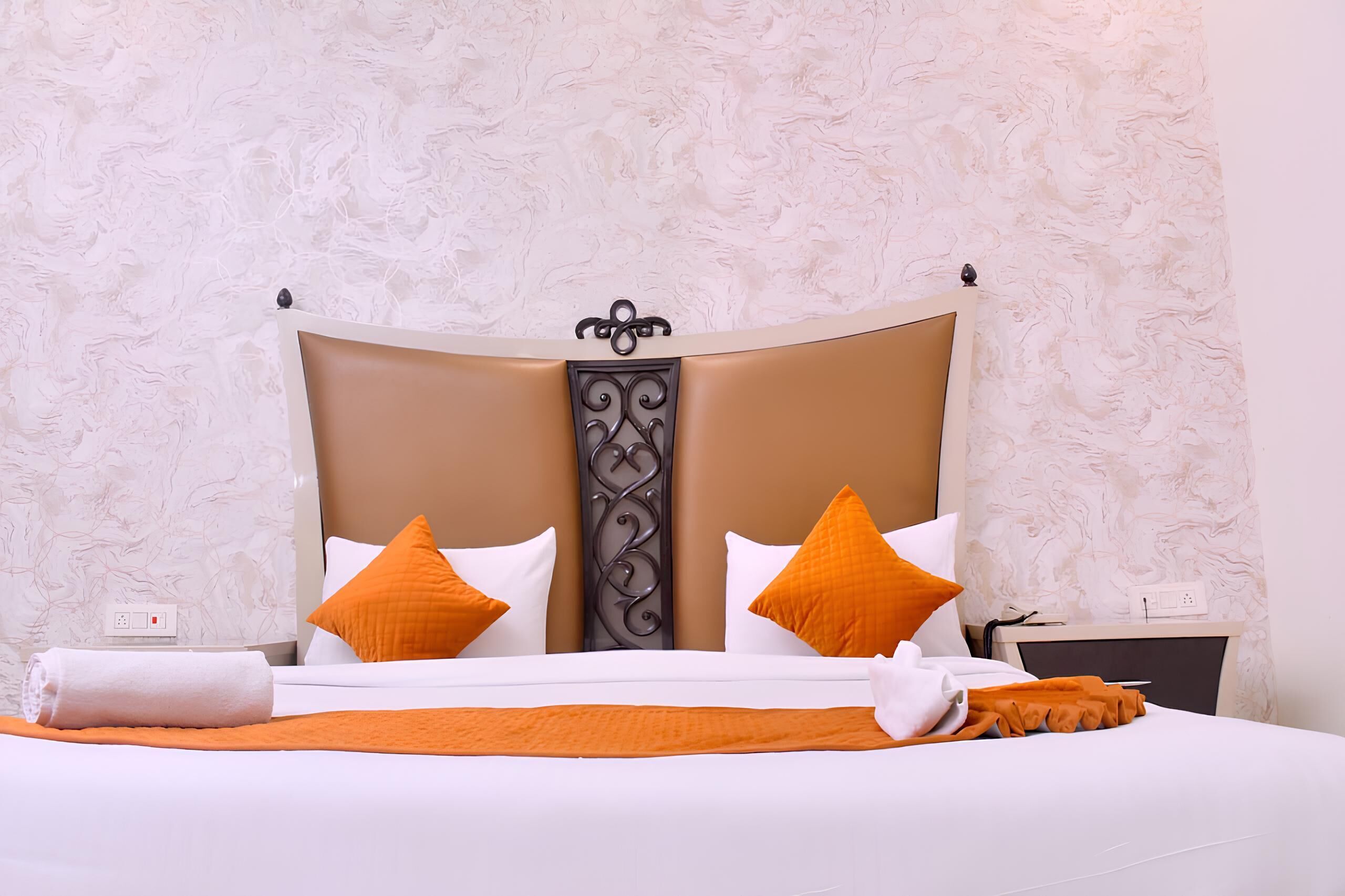 Una cama bien hecha con cabecera marrón, almohadas blancas y anaranjadas, y una manta blanca.