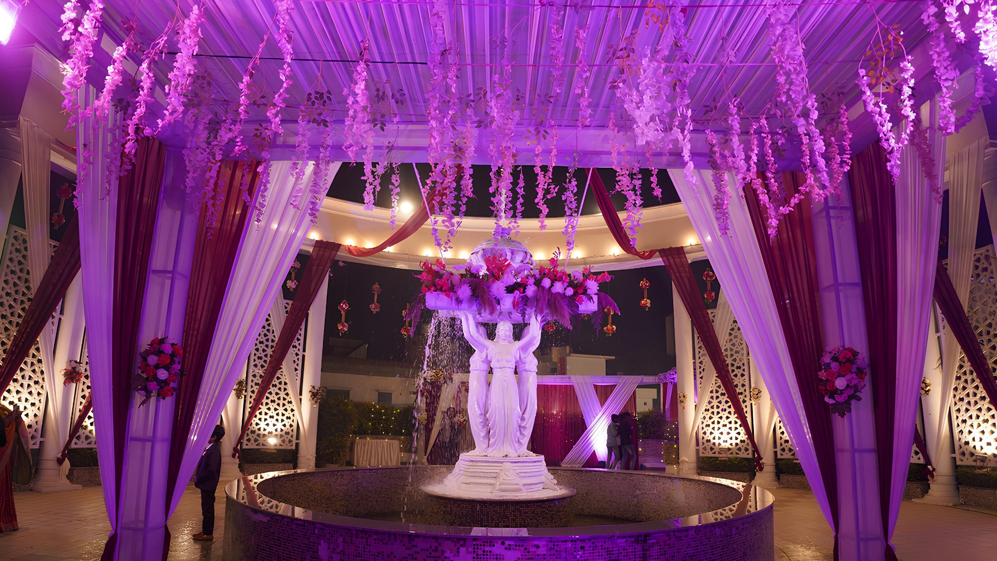 A circular fountain with a statue in the center, surrounded by draped fabric and hanging floral decorations.
