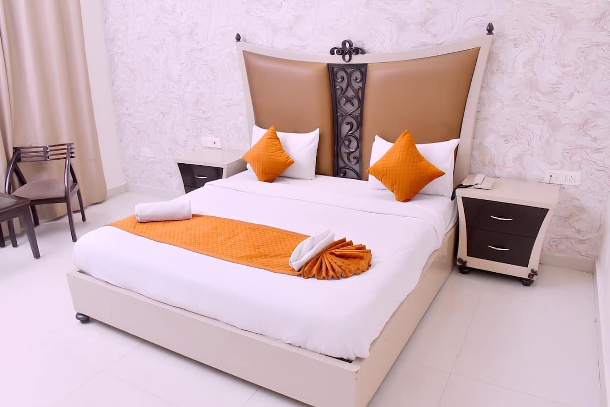 A neatly made bed with a white and orange bedspread, two white pillows, and two orange pillows. There is a small bedside table with a phone and a drawer, and a chair to the left of the bed.