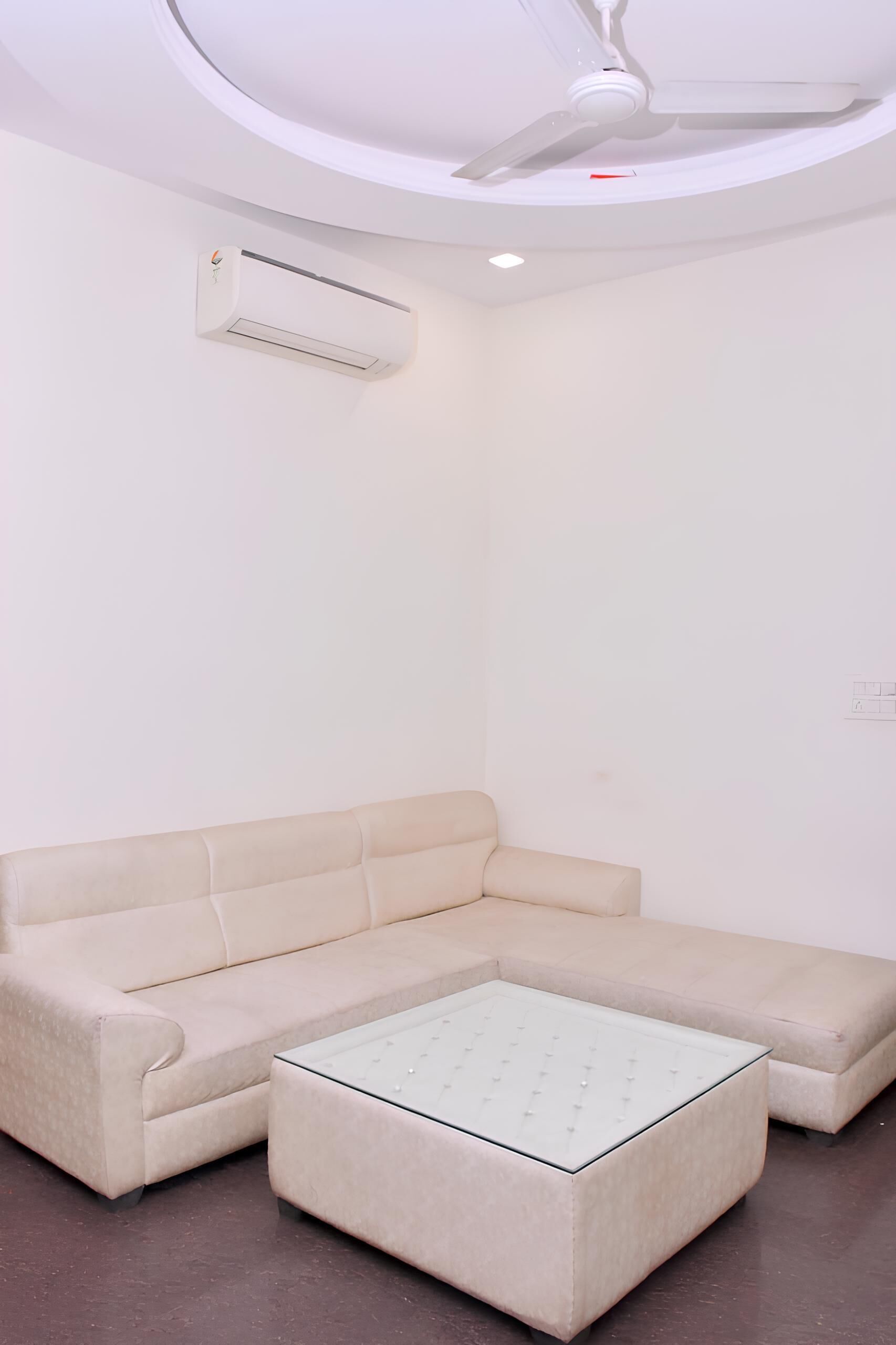 Una habitación moderna y minimalista con un sofá seccional color beige, una mesa de centro de vidrio, un ventilador de techo y un equipo de aire acondicionado.