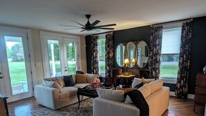 A spacious living room with a large sofa, a coffee table, and a fireplace. There are two windows with curtains, and a ceiling fan is visible.