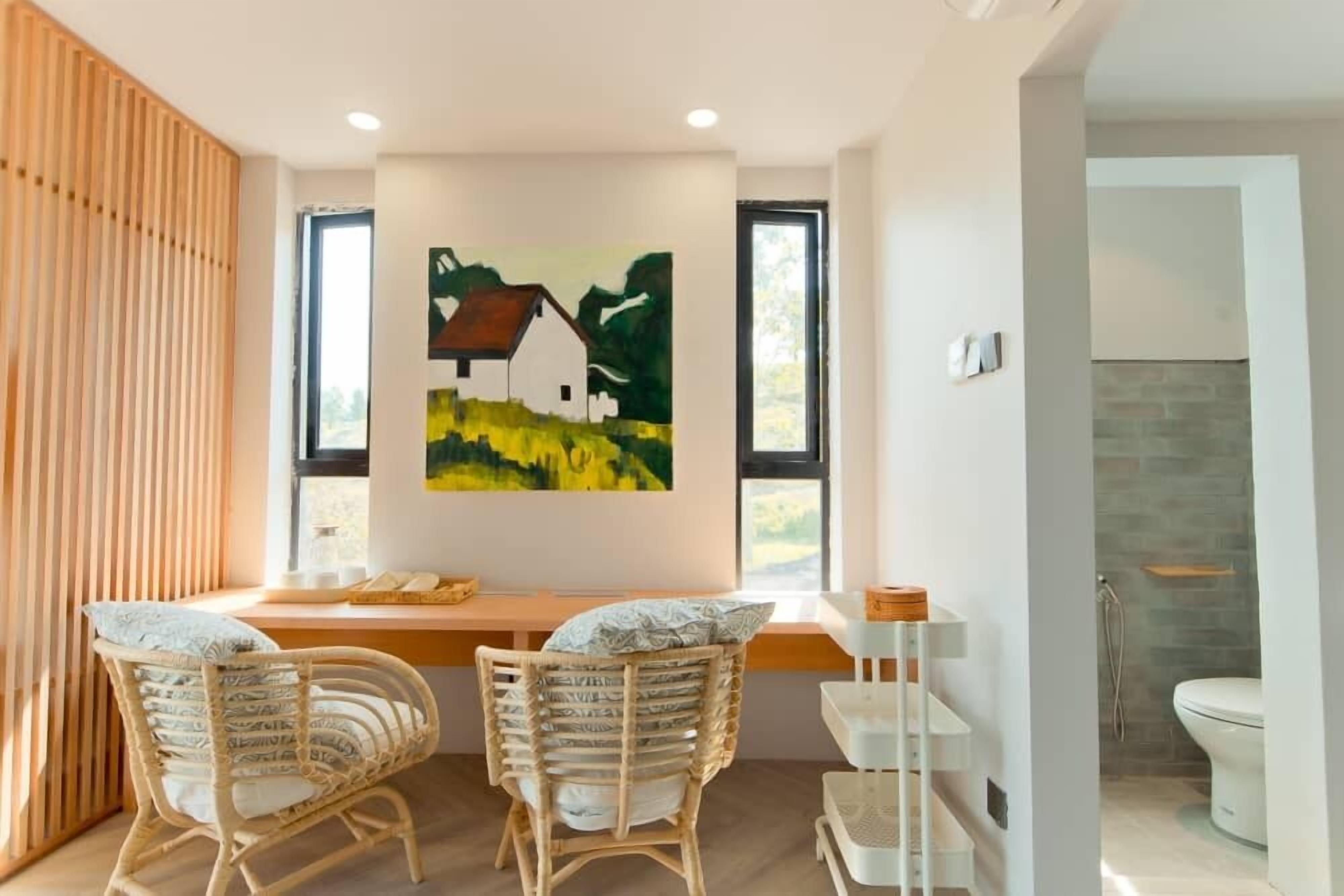Una habitación moderna con un revestimiento de listones de madera, un cuadro de una casa, dos sillas de mimbre, una mesita y vistas a un baño con ducha e inodoro.