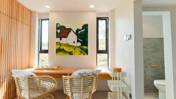 Una habitación moderna con un revestimiento de listones de madera, un cuadro de una casa, dos sillas de mimbre, una mesita y vistas a un baño con ducha e inodoro.