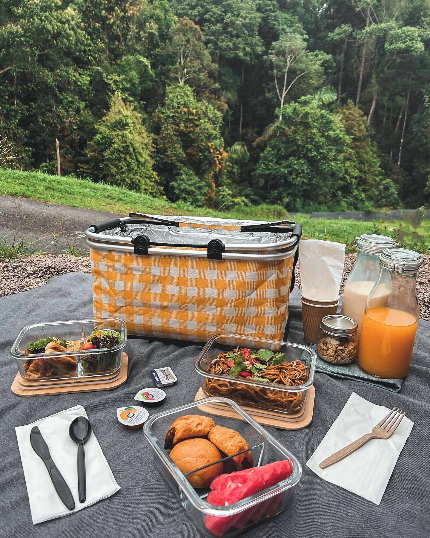 A picnic setup with a checkered bag, food containers, utensils, and a bottle of juice on a blanket in a forested area.