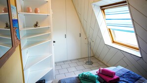Ein Schlafzimmer mit schrägem Dach, einem Oberlicht, einem Bett mit blau-violetter Bettdecke und einem Regal mit Dekorationen.