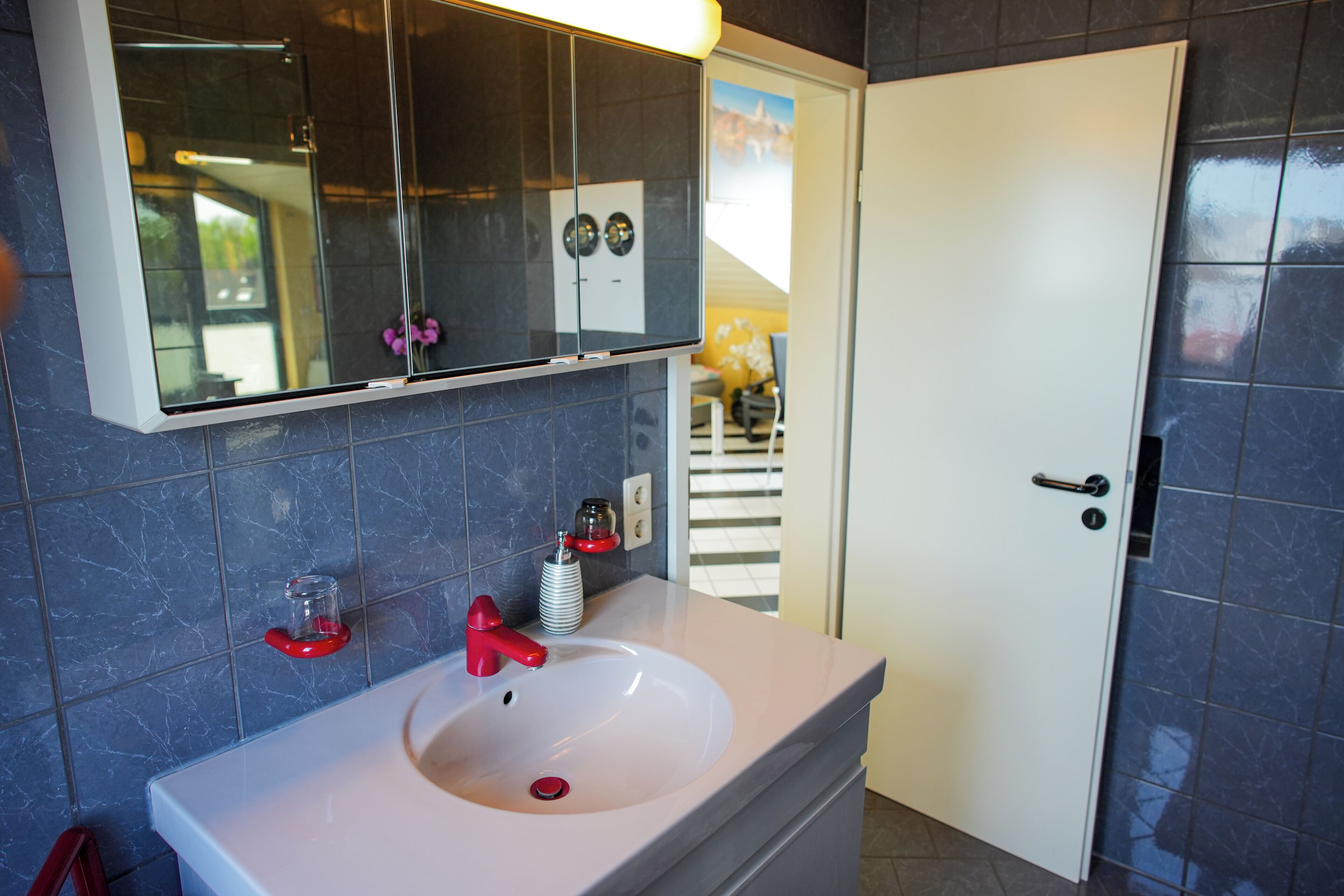 Une salle de bain avec un lavabo blanc, un robinet rouge et un miroir au-dessus. Il y a une porte sur la droite et une fenêtre à l’arrière-plan.