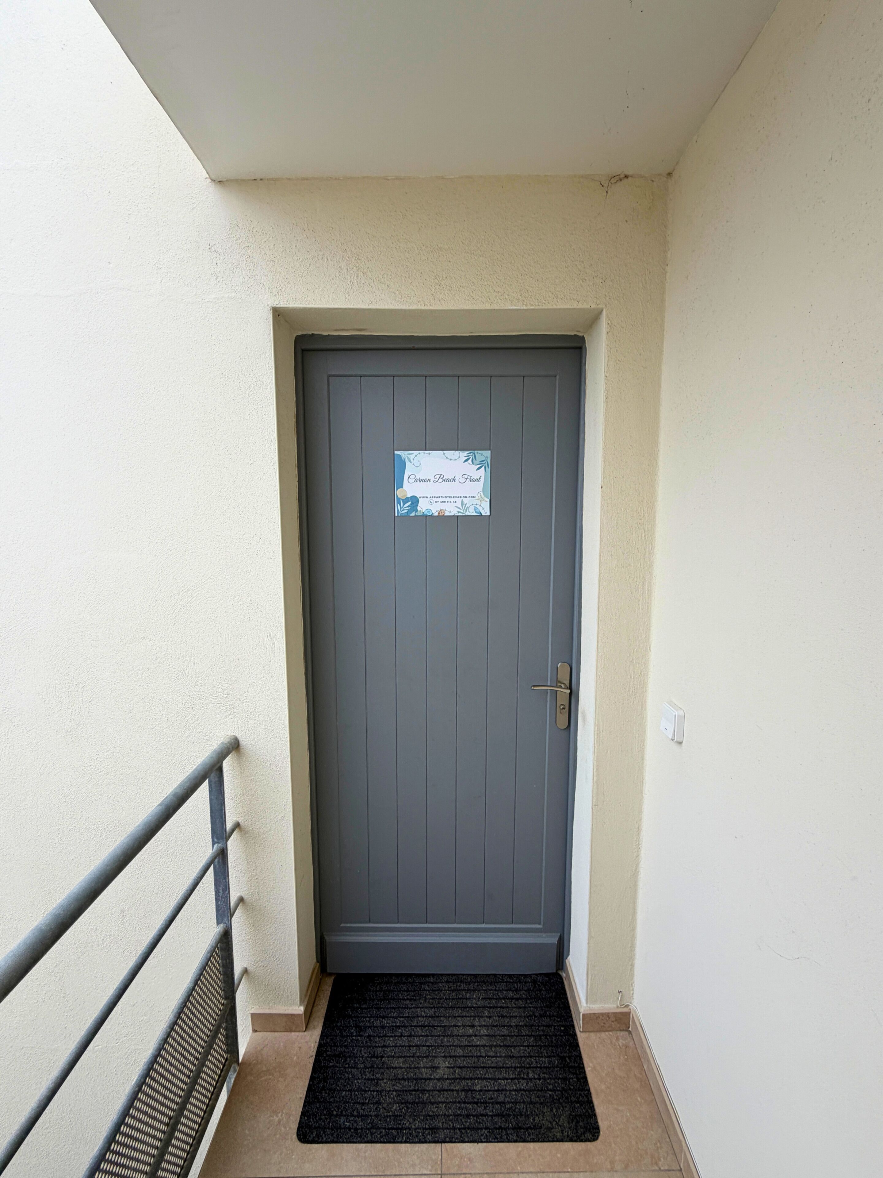Une porte grise avec une plaque, un paillasson noir et une rambarde en métal.