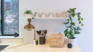 A kitchen with a window, a shelf with kitchenware, a cutting board, a basket, and a potted plant.