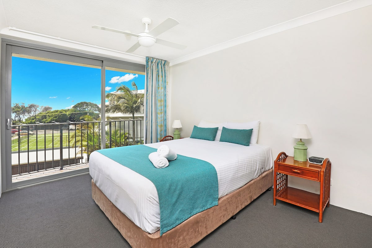 A hotel room with a large bed, a nightstand, a ceiling fan, and a view of the outdoors.