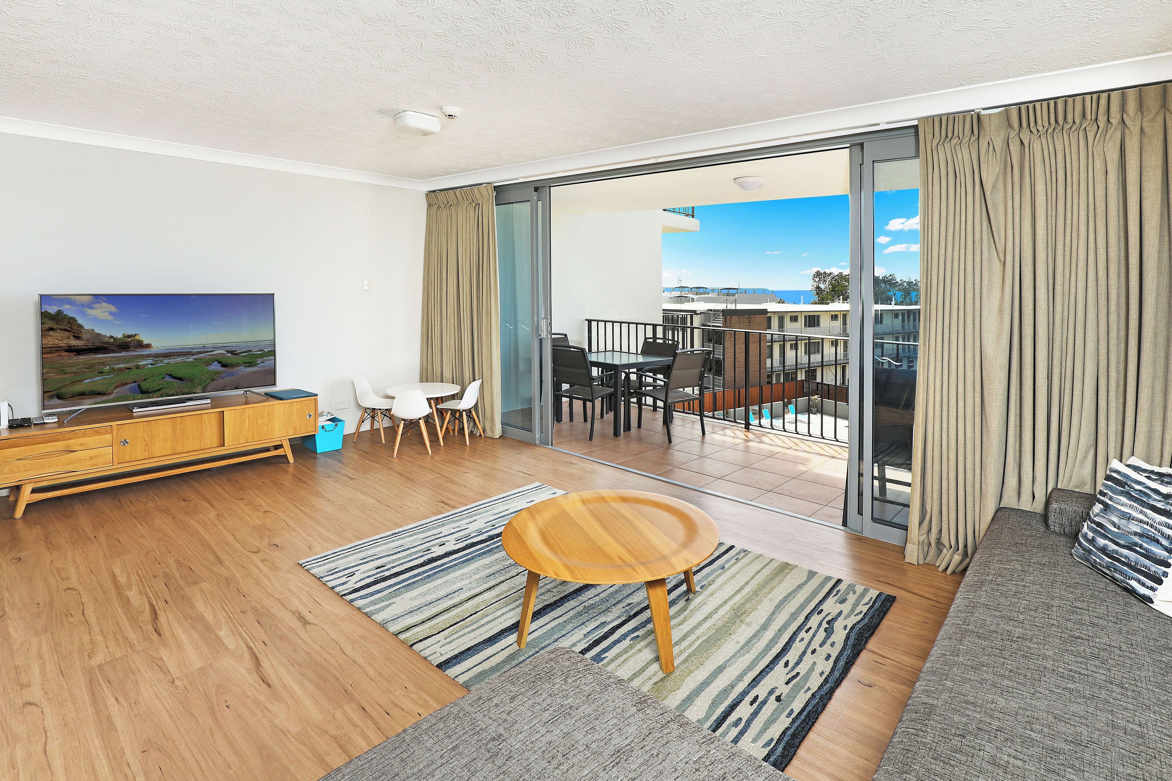 A modern living room with a wooden floor, a round wooden coffee table, a sofa, a TV, and a balcony with a view of the sea.