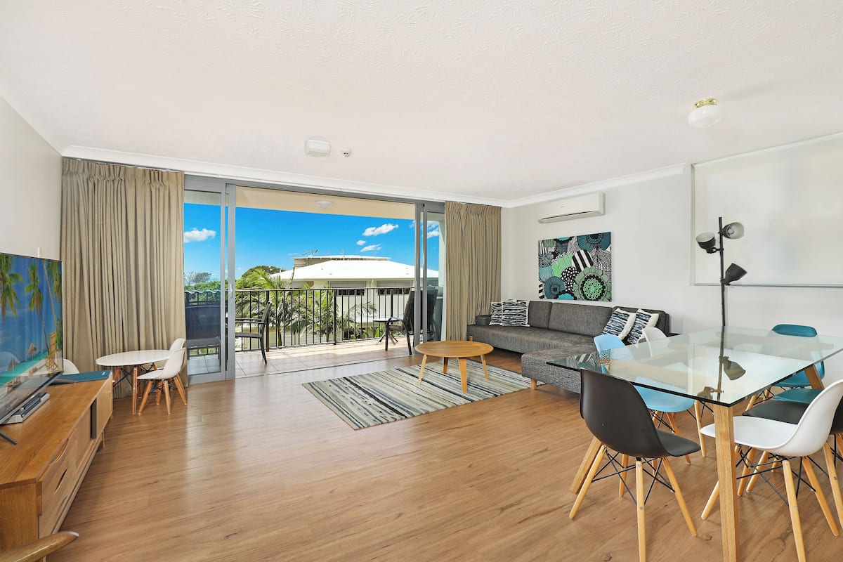 A modern living room with a glass dining table, wooden flooring, and a balcony with a view of the sky and greenery.