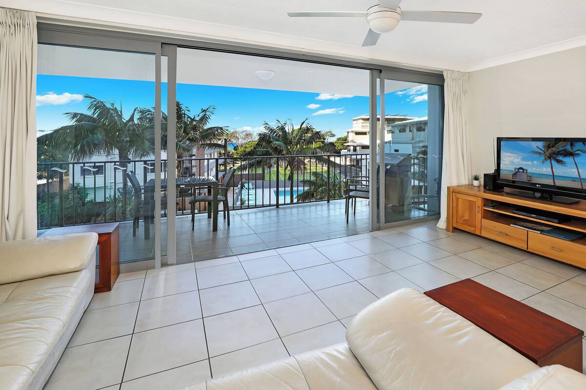A spacious living room with a large sliding glass door leading to a balcony with a pool and palm trees.