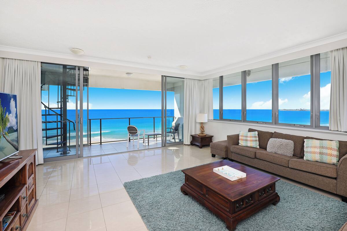A spacious living room with a large window offering a view of the ocean, a wooden coffee table, a sofa with colorful cushions, and a television on a wooden stand.