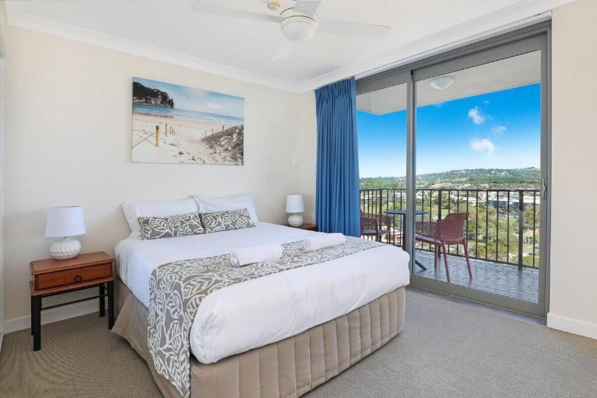 A bedroom with a large bed, a nightstand, a lamp, a painting of a beach, and a balcony with a chair.