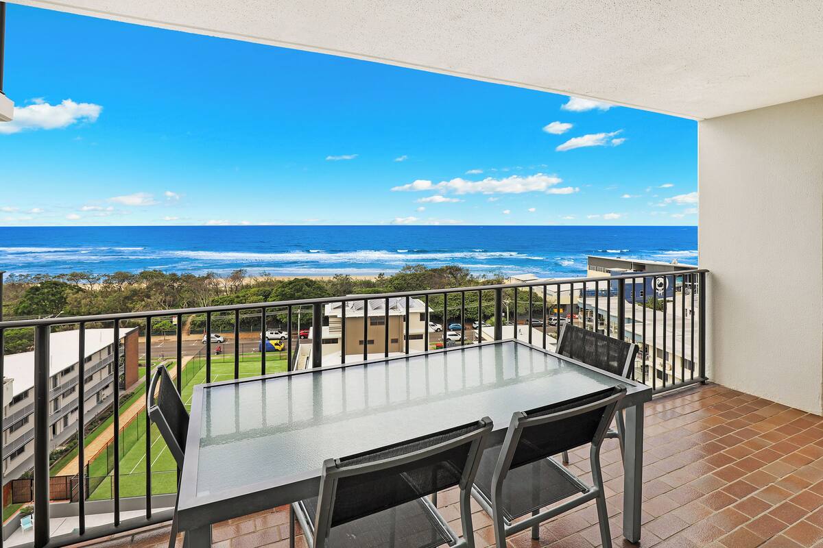 A balcony with a glass table and chairs, overlooking a coastal view with the ocean and a parking area.