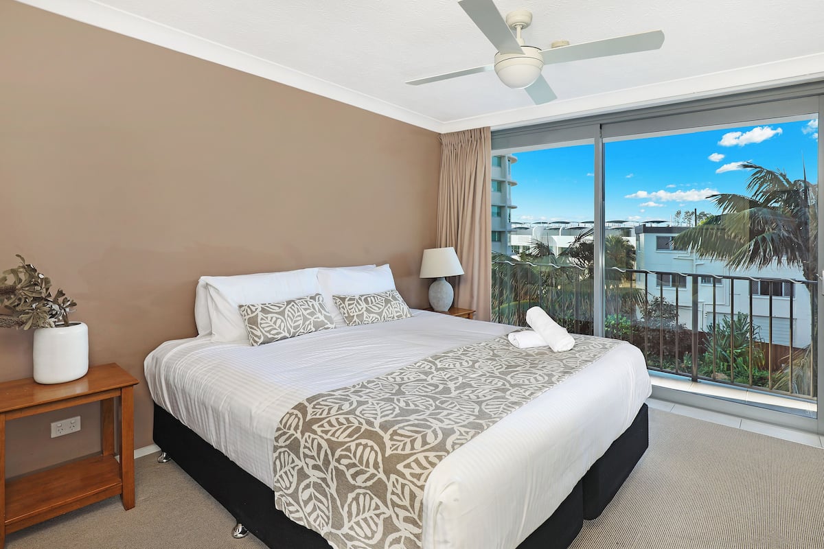 A hotel room with a large bed, a ceiling fan, a nightstand with a lamp, and a view of the city through large windows.
