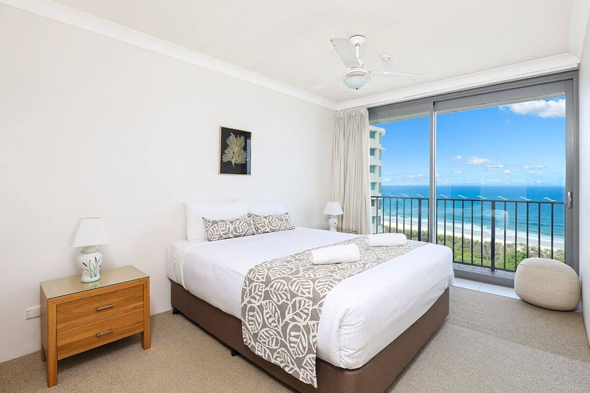 A bedroom with a large bed, a nightstand, a lamp, a framed picture, and a view of the ocean through a balcony.