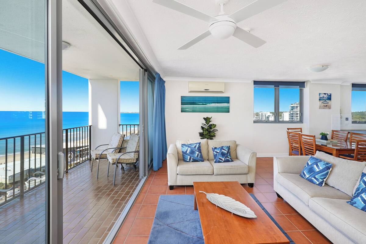 A spacious living area with a balcony overlooking the sea, a ceiling fan, and a dining area with chairs and a table.