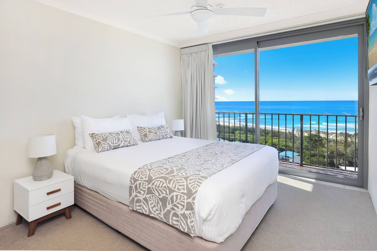 A bedroom with a large bed, a nightstand, a lamp, and a view of the ocean.
