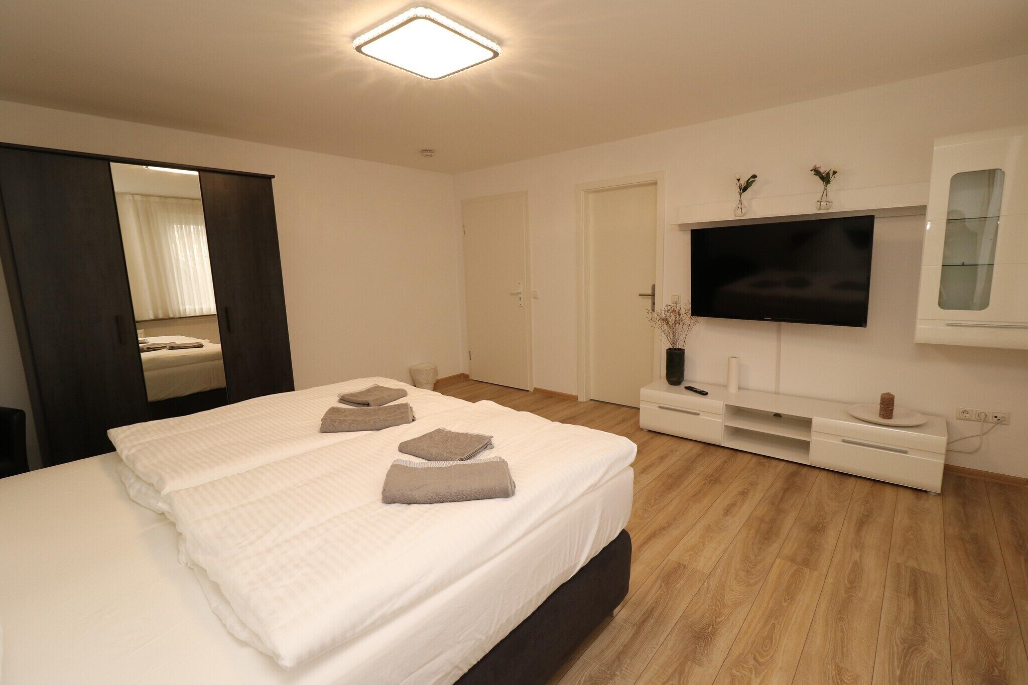 Une chambre d’hôtel moderne avec un grand lit, un téléviseur à écran plat fixé au mur, un plancher en bois et une armoire de couleur sombre.