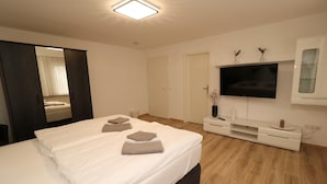 Una habitación de hotel moderna con una cama grande, un televisor de pantalla plana montado en la pared, piso de madera y un armario de color oscuro.