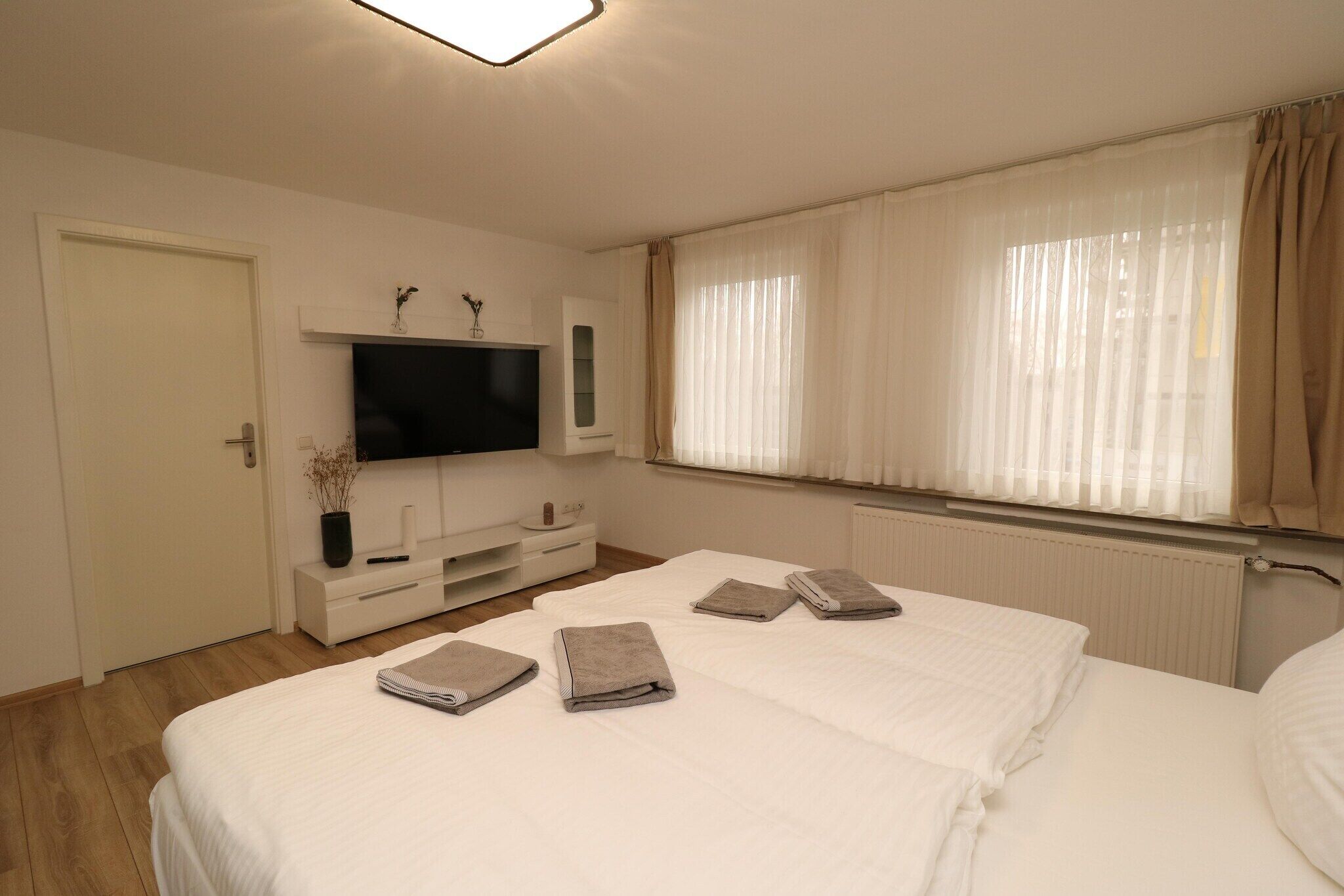 Quarto de hotel com cama, televisão, janela com cortinas, mesa de cabeceira e radiador.