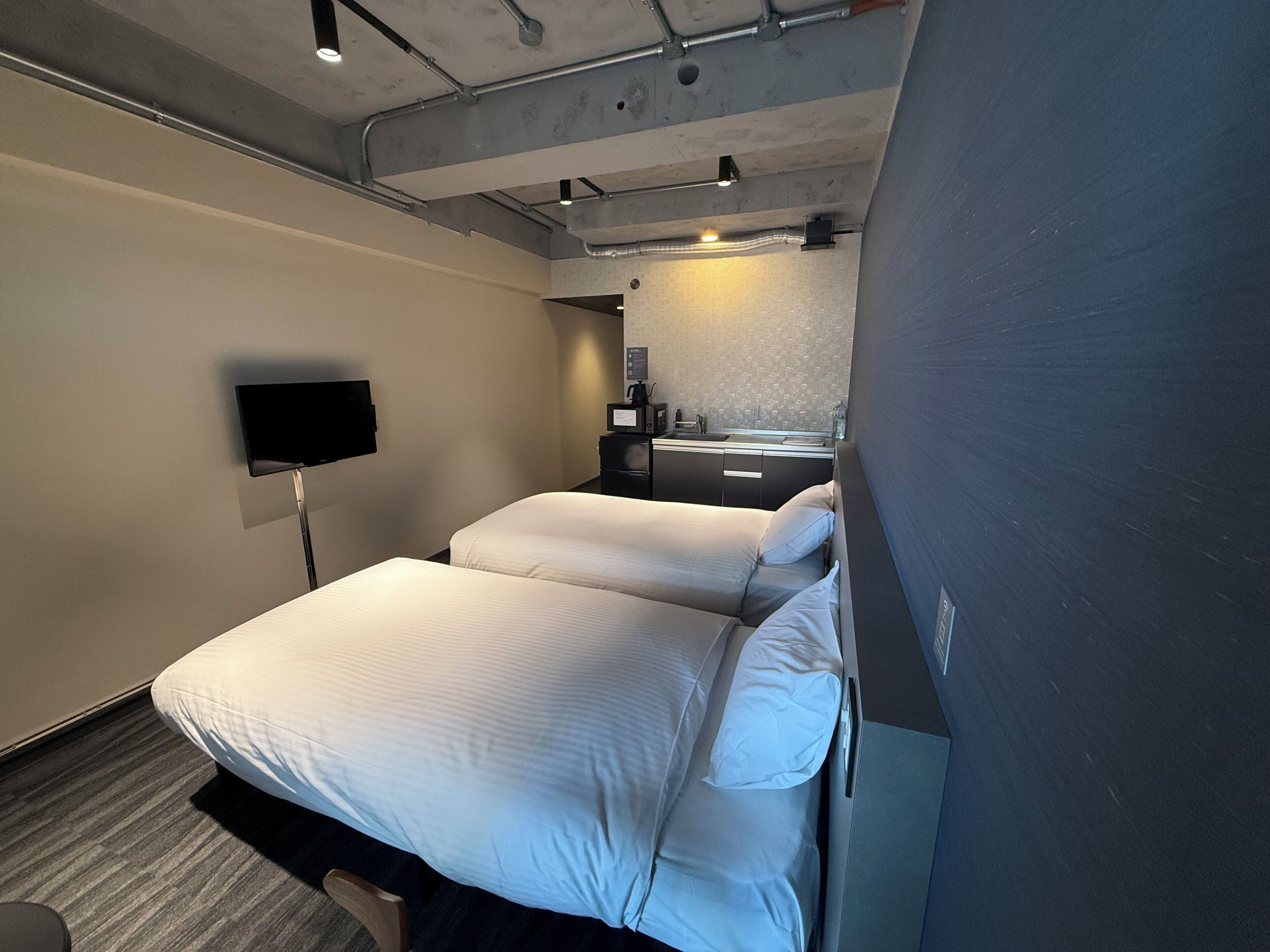 A hotel room with two beds, a flat-screen TV, a small desk with a mirror, and a kitchenette.