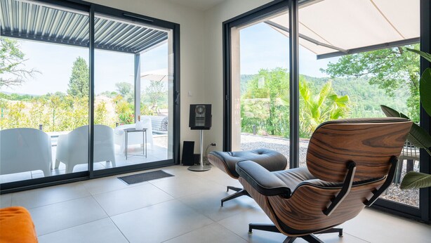 A modern interior with large windows, a ceiling fan, and a view of a garden. - Villa Port des Vins – Luxury villa in Salernes with heated pool (Salernes)