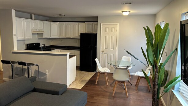 Un espacio habitable compacto con kitchenette, un comedor con mesa y sillas de vidrio, un sofá y una planta en maceta.