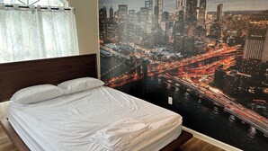 Un dormitorio con un gran mural de paisaje urbano, una cama con ropa de cama blanca, un cabecero de madera y una ventana con cortinas traslúcidas.