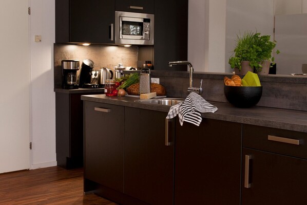 A modern kitchen with dark cabinetry, a stainless steel sink, a microwave, and a countertop with a coffee maker, toaster, and a bowl of fruit.