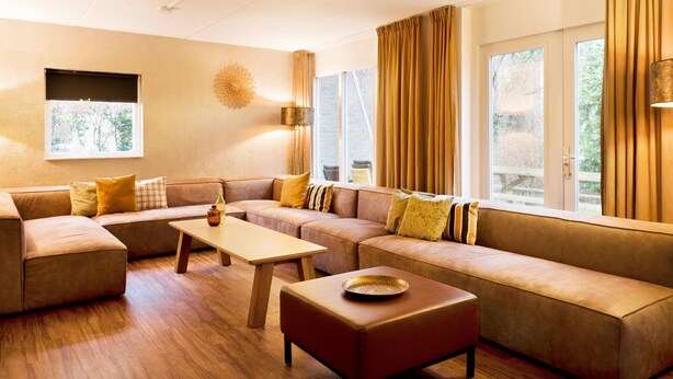 Una sala de estar moderna con un sofá modular, una mesita de centro y un pequeño otoman. Hay ventanas grandes con cortinas, una lámpara de pared y una lámpara decorativa con forma de sol.