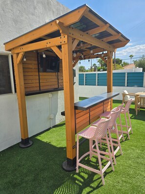Een houten pergola met een bar en zitgedeelte, inclusief een televisie en buitenverlichting.