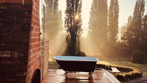 Une terrasse en bois avec une baignoire, entourée d’un mur de briques et de grands arbres.