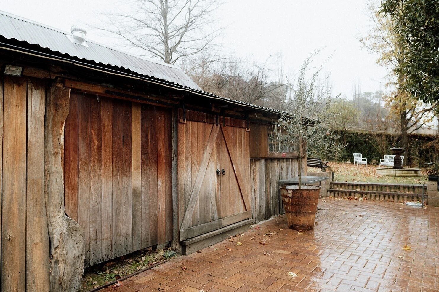 A rustic wooden building with a metal roof, a large sliding door, and a brick patio area with benches and a small fountain.
